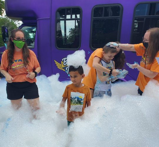 Staff in orange shirts and children play in huge piles of foamy bubbles