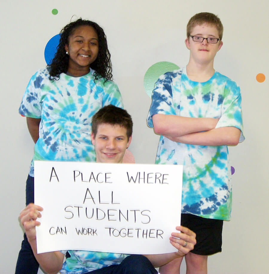 Three smiling children wear matching tie dye shirts and one holds posterboard that says "a place where all students can work together"
