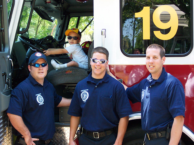 Three firefighters smile while young child sits in driver's seat of firetruck